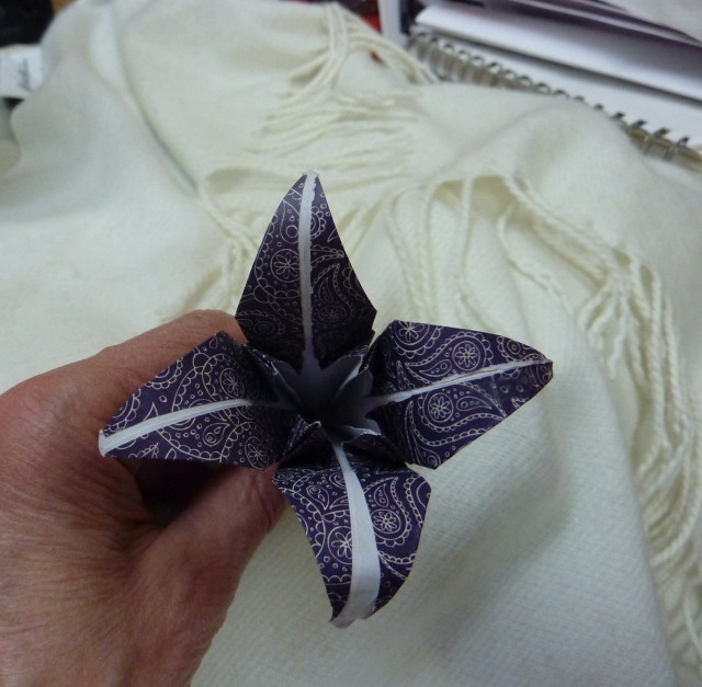 An origami flower made from purple paisley paper; it has four petals curving outward. 
