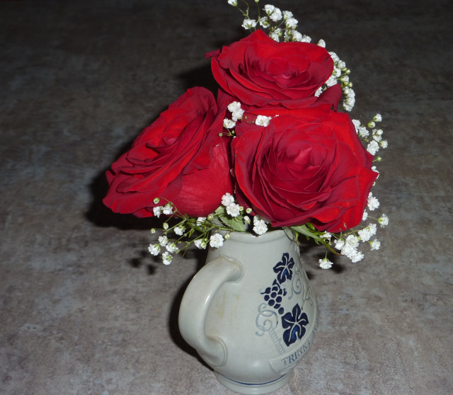 Three dark red roses are tucked into a tiny grey pottery pitcher. Some baby's breath flowers as well.