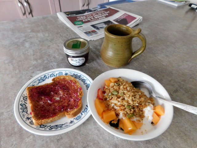 Photo of breakfast: a bowl of fresh fruit, yogurt and granola; a slice of toast spread with jam, a mug of coffee, and a small jar of homemade jam.